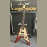 Elektrická kytara s červeným tělem ve tvaru V a krčkem z tmavého dřeva. Na bílém pickguardu jsou dva humbuckery a tři ovládací knofíky. Kytara je opř…