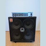 Světle modrý zesilovač značky Hartke, model MODEL HA3000 - 350 WATTS, umístěný na černém basovém reproduktoru. Reproduktor má čtyři membrány kryté ko…