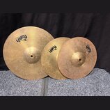 Tři mosazné činely různých velikostí s viditelným povrchovým opotřebením a šmouhama. Každý činel nese logo „flash CYMBALS“. Na okraji největšího čine…