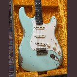 Světle modrá elektrická kytara typu Stratocaster s bílým pickguardem a třemi snímači. Tělo vykazuje výrazné opotřebení laku, zejména v dolní části a…