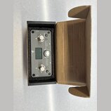 Černý elektronický přístroj v kartonové krabici. Na předním panelu jsou dva kovové konektory, tři stříbrná tlačítka a malý displej. Jsou zde nápisy U…
