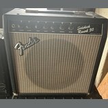 Kytarový kombo zesilovač v černém obalu s hnědou textilní maskou. Na horním ovládacím panelu jsou nápisy Sidekick Reverb 30 a Fender Musical Instrume…
