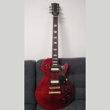 Červená elektrická kytara s dumpedým topem a černým pickguardem. Na hlavě hmatníku je nápis Epiphone. Kytara má dva humbuckery se zlatými kryty, čtyř…