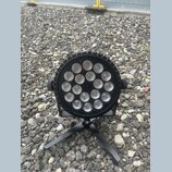 Černý kulatý LED reflektor s 18 svítícími elementy v mřížce. Zařízení je umístěno na černém trojnožném stojanu a má po stranách dvě ovládací knoflíky…