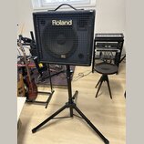 Černý kombozesilovač značky Roland s nápisem KC na spodní části mřížky. Zařízení je umístěno na černém kovovém stativu s trojnožkou. V pozadí jsou vi…