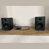 Dva černé studiové monitory s kulatými membránami v zelených rámečcích. Na spodní části obou reproduktorů je bílé kulaté logo s písmenem G. Mezi repr…