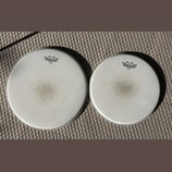 Dvě bílé bubnové kožehy značky REMO. Obě mají v horní části logo s nápisem REMO a pod ním text EMPEROR COATED MADE IN U.S.A. Povrchy kožehů vykazují…