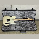 Elektrická kytara typu Telecaster v krémové barvě s bílým pickguardem, uložená v černém tvrdém kufru s šedou polstrováním. Na hlavě hmatníku je vidit…
