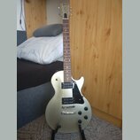 Stříbrná elektrická kytara s černým pickguardem a dvěma humbuckery. Na hlavě hmatníku je viditelné logo Gibson. Nástroj je umístěn ve stojanu v inter…