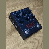 Černý kytarový pedál s nápisy TRIO+ a Band Creator+ Looper. Zařízení obsahuje několik ovládacích knoflíků, přepínačů a sekci s označením GENRE, STYLE…
