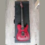 Červená elektrická kytara s matným povrchem položená na černém tvrdém kufru. Na hlavě hmatníku je bílý nápis "Schecter". Nástroj má dva humbuckery, j…