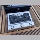 Stříbrný kytarový předzesilovač s nápisy AMP 1 a BluGuitar TONE INNOVATION. Na panelu jsou ovládací prvky s popisky VOLUME, GAIN, MASTER, BASS, MIDDL…