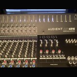 Fotografie zachycuje část míchacího pultu s nápisem AUDIENT ASP 4816 HERITAGE EDITION. Zařízení má šedý kovový povrch a je vybaveno řadou posuvníků,…
