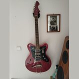 Červená elektrická kytara s polodupatým korpusem a f-otvory, zavěšená na bílé stěně. Nástroj má tmavý hmatník, černý pickguard a stříbrný vibrační sy…