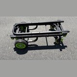 Černý transportní vozík s kovovou konstrukcí a zelenými detaily. Má dvě velká pneumatická kola vpředu a dvě menší otočná kolečka vzadu. Na zelených p…