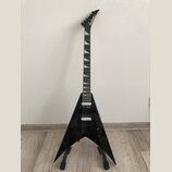 Černá elektrická kytara s tělem ve tvaru V umístěná na stojanu. Na hlavě hmatníku je bílý nápis Jackson. Kytara má dva humbuckery, jeden ovládací kno…