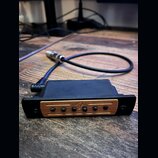 Černý kabel s konektorem, který je připojen k malému černému boxu. Na přední straně boxu je mosazný nebo zlatý kovový panel s pěti malými černými kno…