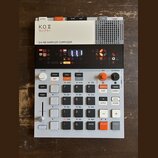 Bílo-šedý sampler s černým displejem a oranžovými detaily. Na horním bílém panelu je nápis K.O.II, pod ním japonské znaky サンプラー a text 64 MB SAMPLER…