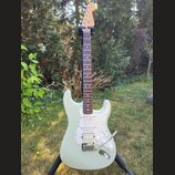 Světle zelená elektrická kytara typu Stratocaster s bílým perleťovým pickguardem a hnedým hmatníkem. Na hlavě je vidět nápis Fender. Nástroj stojí na…