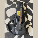 Elektrická kytara s matně černým povrchem, černým hardwarem a hnedým hmatníkem. Na hlavě je bílý nápis Ibanez a na černém obalu v pozadí je bílé logo…