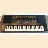 Černý digitální klavír s bílými a černými klávesami. Na ovládacím panelu je nápis YAMAHA PSS-590 a logo AWM. Přístroj obsahuje červené digitální disp…
