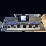 Stříbrný digitální klavír s nápisy YAMAHA a Tyros 5. Nástroj má v horní části displej, na kterém je zobrazen text Tyros 5 a další systémové informace…