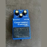 Modrý efekt pedál s nápisy LEVEL, SUSTAIN, OUTPUT a INPUT. Na přední straně je bílý text Compression Sustainer CS-2 a černé logo BOSS. Zařízení vykaz…