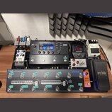 Sestava efektů na černém pedalboardu. V horní části jsou umístěny jednotky Fractal Audio AM4, Shure s displejem a bílý pedál Noise Suppressor NS-2. V…