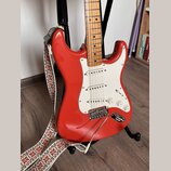 Červená elektrická kytara typu Stratocaster s bílým pickguardem a světle hnědým javorovým hmatníkem. Nástroj má tři snímače, tři ovládací knofíky a c…