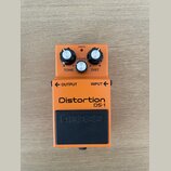 Oranžový efekt pedál s černým stoupacím spínačem. Na zařízení jsou nápisy Distortion DS-1, BOSS, LEVEL, TONE, DIST, CHECK, OUTPUT a INPUT. Povrch ped…
