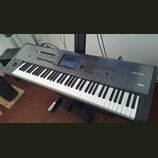 Šedý digitální klavír s černými a bílými klávesami, umístěný na černém stojanu. Na pravé straně jsou bílé nápisy KRONOS MUSIC WORKSTATION a KORG. Nás…