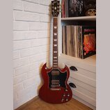 Tmavě červená elektrická kytara s černým pickguardem a hnědým hmatníkem. Na hlavě je nápis Gibson a označení SG, na hmatníku jsou vidět trapezoidní v…