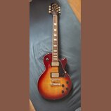 Elektrická kytara s trupem v červeno-žlutém sunburst provedení a hnědým hmatníkem s trapezoidními vložkami. Na hlavě je nápis Gibson, modelové označe…