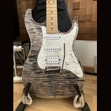 Elektrická kytara typu Stratocaster s tělem z vyvzorovaného dřeva v šedohnědé barvě a hmatníkem ze světlého javoru. Nástroj má bílý pickguard s perle…