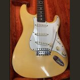 Krémově bílá elektrická kytara typu Stratocaster s bílým pickguardem a hnedým hmatníkem. Na kytaru jsou instalovány tři snímače s bílými plastovými k…