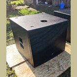 Černý subwoofer v kostkovém provedení s texturovaným povrchem. Přední strana je kryta kovovou ochrannou mřížkou, na které je umístěno malé logo JBL.…