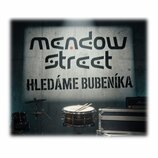 Fotografie zobrazuje buben s položenými paličkami, část činelu na stojanu a černý transportní case. V pozadí je zeď s velkým nápisem „meadow street“…