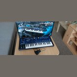 Modrý kompaktní syntetizátor s bílými klávesami a černými ovládacími prvky. Na přístroji je nápis MININOVA 2 a logo novation. Syntetizátor stojí před…