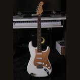 Bílá elektrická kytara typu Stratocaster s hnědým pickguardem a hnedým hmatníkem. Na hlavě je vidět nápis Fender. Kytara má tři snímače, tři ovládací…