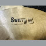 Detailní pohled na mosazný činel s viditelným broušením povrchu. Na čineli je černý nápis SWEET HI HAT a pod ním text 15"/38 cm TOP. Povrch vykazuje…