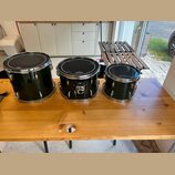 Tři černé bubny s kovovými obručemi a ladidly umístěné na dřevěném stole. Na každém z bubnů je v horní části bílý nápis "Gretsch". Povrch nástrojů je…