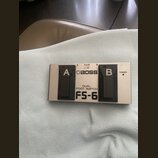Stříbrný dvoupedálový spínač s černými plasty. Na horní straně je logo BOSS a nápisy DUAL FOOT SWITCH oraz FS-6. Pedály jsou označeny písmeny A a B,…