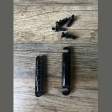 Na fotografii jsou dvě černé kovové součástky a pět černých šroubů. Jedna součástka je úzký profilovaný pás s otvory na koncích, druhá je hladký vále…