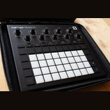 Černý sekvenční synthesizer Novation Circuit Tracks v černém přenosném pouzdře. Horní část obsahuje několik otočných ovladačů s bílými popisky jako M…