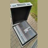 Otevřený hliníkový transportní kufr s černým polstrováním uvnitř, ve kterém je umístěn šedý mixážní pult. Na přístroji jsou vidět četné posuvníky, ot…