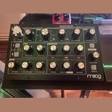 Černý analogový basový syntetizátor s nápisem MINITAUR a analog bass synthesizer. V levém dolním rohu je bílé logo moog. Přístroj obsahuje několik řa…