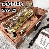 Zlatý altový saxofon v hnědém tvrdém kufru s polstrováním. Na obrázku je text YAMAHA YAS-23 a v kufru se nachází také munční dutina, hnedbek s rýčkem…