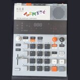 Šedý sampler s názvy K.O.II a nápisem サンプラー, pod kterým je text 128 MB SAMPLER COMPOSER. Zařízení obsahuje digitální displej, několik tlačítek v bílé…