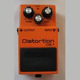 Oranžový efekt pedál s nápisy Distortion a DS-1. Na horní části jsou tři ovládací knoflíky s popisky TONE, LEVEL a DIST a červená kontrolka CHECK. Ve…
