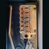 Zlatý Tremolo bridge pro elektrickou kytaru s šestnácti sedly a součástí je páka. Kovový povrch vykazuje známky opotřebení, drobné škrábance a oxidac…
